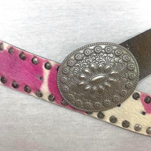 Op de afbeelding: Een bruine leren riem met een grote zilveren gesp met een bloemenmotief. De riem is versierd met roze en wit koehuid en zilveren studs.