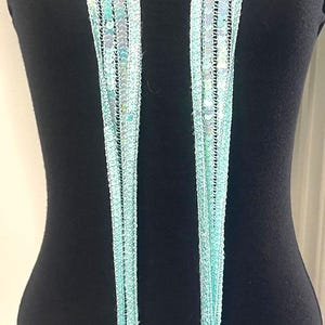 May include: A light blue scarf with a sequined design and fringe. The scarf is displayed on a black mannequin. The scarf is made of a delicate, open weave material.