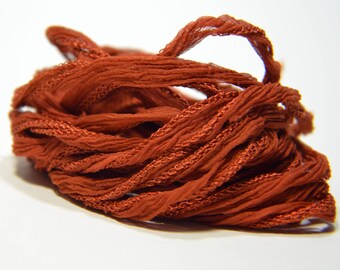 May include: A close-up of a spool of rust-colored silk ribbon. The ribbon is thin and has a slightly textured surface.