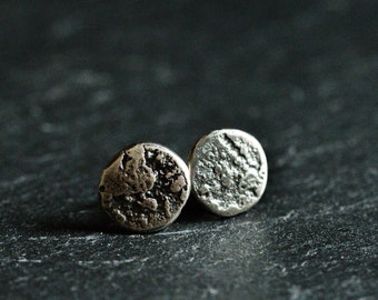 May include: Two small, round silver stud earrings with a textured surface. The earrings have an irregular, organic design, giving them a handcrafted look. They are presented on a dark, textured background.