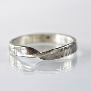 Op de afbeelding: Een zilveren ring met een enkele draai in de band. Het metalen oppervlak heeft een getextureerde, geborstelde afwerking met zichtbare verticale strepen over de gehele omtrek, wat zorgt voor een matte uitstraling met subtiele accenten.