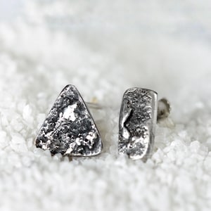 Puede incluir: Un par de pendientes de plata. Uno es triangular y el otro rectangular. Ambos tienen un aspecto texturizado y martillado. Los pendientes se muestran sobre una superficie blanca texturizada.