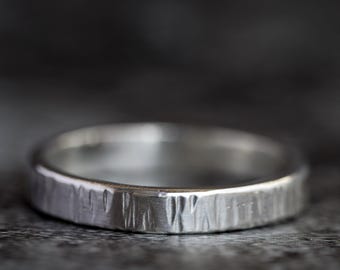 May include: A silver ring with a textured, bark-like surface. The band is a simple, flat design, with a slightly worn appearance. The ring is set against a dark, blurred background, highlighting its metallic sheen.