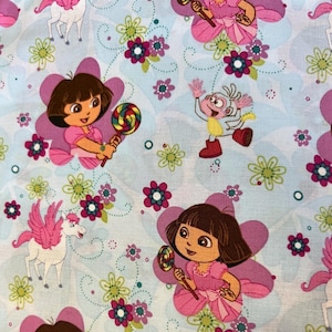 May include: Light blue fabric featuring cartoon characters Dora the Explorer and Boots, surrounded by pink flowers, green flowers, and white winged horses. Dora is holding a colorful lollipop. The fabric has a whimsical, playful design.