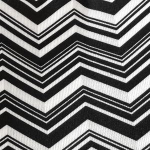 May include: Close-up of a fabric with a black and white chevron pattern. The design features bold, zigzagging lines that create a dynamic visual effect. The fabric has a textured appearance, suggesting a woven material.