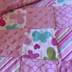 May include: A patchwork quilt featuring squares of pink and white fabrics. Some squares have a textured, dotted pattern, while others display butterfly and floral designs in shades of pink, green, and blue. Diagonal stripes and a soft, plush edge complete the design.