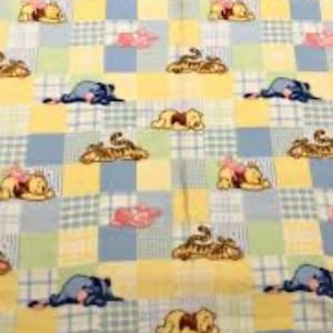 May include: A baby blanket featuring a patchwork design with Winnie the Pooh, Tigger, and Eeyore characters. The squares are yellow, blue, and green, with pink accents. The blanket is likely made of soft fabric, suitable for a nursery.