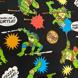 May include: Black fabric featuring a repeating pattern of green Teenage Mutant Ninja Turtles in action poses. Comic book style speech bubbles with phrases like "Tough as a Turtle!" and "Pizza, Please!" are scattered throughout.