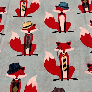 May include: Light blue fabric with a repeating pattern of red foxes. Each fox is dressed in a vest, tie, or hat, and some wear monocles. The foxes have white-tipped tails and are set against a light blue background.