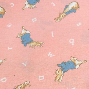 May include: A light pink fabric featuring a repeating pattern of brown rabbits wearing blue jackets, interspersed with scattered white lowercase letters of the alphabet. The rabbits are depicted in various orientations across the soft-textured material.