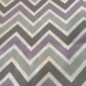 May include: A fabric with a repeating chevron pattern. The design features alternating bands of white, light gray, medium gray, and lavender. The zig-zag pattern creates a visually dynamic and modern aesthetic.