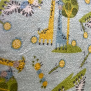 May include: Light blue fabric with a repeating pattern of yellow and blue giraffes, green trees, and stylized flowers. The giraffes have yellow bodies and brown spots. The trees are green with yellow trunks. The flowers are yellow with white centers.