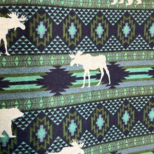 May include: A patterned fabric featuring a repeating design of white moose and bear silhouettes against a backdrop of geometric shapes in shades of blue, teal, and navy. The fabric has a textured appearance.