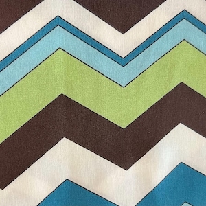 May include: A close-up of a fabric with a repeating chevron pattern. The pattern features alternating zigzags in brown, white, light blue, and green. The fabric has a slightly textured appearance.