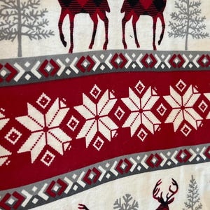 May include: A white fabric with a repeating pattern of red and black plaid deer, gray trees, and snowflakes. The design includes red bands with white snowflake patterns and gray geometric borders, creating a festive winter theme.