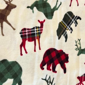 May include: Cream-colored fabric featuring a repeating pattern of woodland animals, including deer and bears. The animals are filled with plaid patterns in red, green, and brown, creating a rustic and outdoorsy theme.