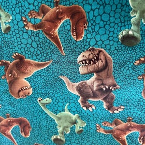 May include: A teal fabric with a repeating pattern of cartoon dinosaurs in shades of brown and green. The dinosaurs are in various poses, with some showing their teeth. The background has a textured, scale-like design.