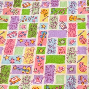 May include: A fabric print featuring a repeating pattern of small cartoon dragons in various colors, interspersed with musical notes, guitars, paint palettes, paintbrushes, books, and stars, arranged within a grid of light-colored rectangles.