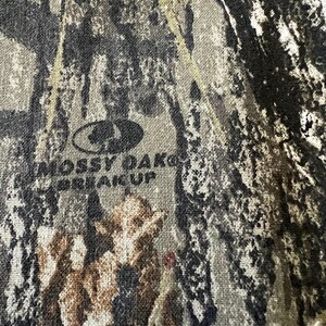 May include: Close-up of a camouflage pattern fabric with the Mossy Oak logo and the words "Break-Up" printed in dark gray. The fabric features a blend of brown, green, and gray tones.