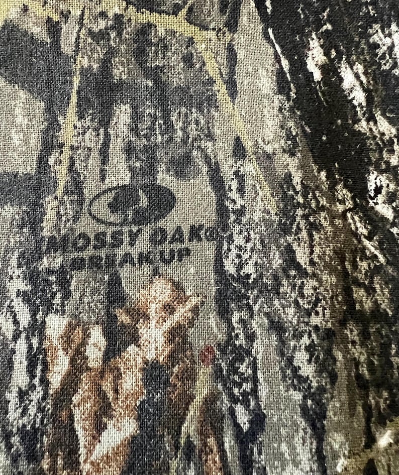 May include: Close-up of a camouflage pattern fabric with the Mossy Oak logo and the words "Break-Up" printed in dark gray. The fabric features a blend of brown, green, and gray tones.