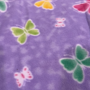 May include: A close-up of a soft, purple fabric with a repeating pattern of butterflies in various colors. The butterflies are outlined in white and filled with shades of green, yellow, and pink. The fabric has a slightly fuzzy texture.