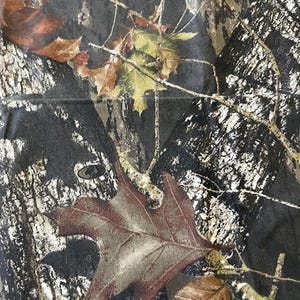 May include: Close-up of a camouflage pattern featuring a blend of brown, green, and black hues. The design incorporates detailed depictions of leaves and tree bark, creating a naturalistic appearance. The fabric has a slightly textured surface.