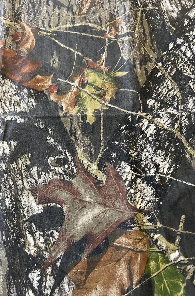May include: Close-up of a camouflage pattern featuring a blend of brown, green, and black hues. The design incorporates detailed depictions of leaves and tree bark, creating a naturalistic appearance. The fabric has a slightly textured surface.