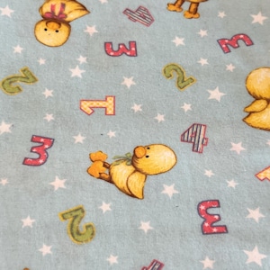 May include: Light blue fabric with a repeating pattern of yellow ducklings, numbers, and white stars. The numbers are in various colors and styles, with the ducklings in different poses. A whimsical design for children's items.