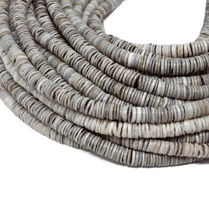 May include: A coiled strand of gray and white shell beads. The beads are flat, disc-shaped, and have a natural, organic appearance. The beads are tightly strung together, creating a textured surface. The color palette is neutral, with variations of gray and white.