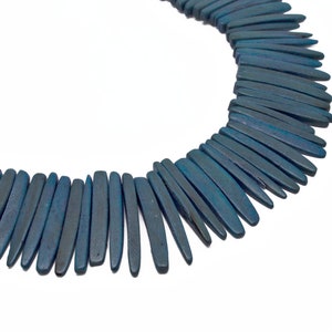 May include: A necklace made of long, thin, teal-colored wooden beads. The beads are arranged in a graduated pattern, with the longest beads in the center and the shortest beads on the edges.