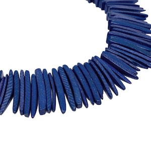 May include: A dark blue necklace composed of numerous, elongated, wooden beads. The beads are arranged in a curved shape, with a textured surface. The necklace is set against a white background, highlighting the color and design of the jewelry.