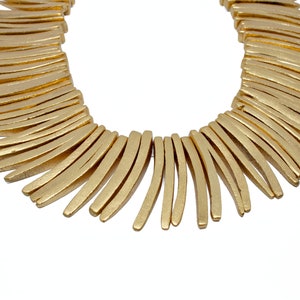May include: A gold-colored necklace with a series of curved, rectangular elements. The necklace has a matte finish and is set against a white background. The design is modern and geometric.