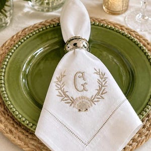 Peut inclure: Une serviette en tissu blanc ornée d'un monogramme beige brodé représentant la lettre G, entourée d'une couronne de laurier et d'un motif de coquillage. La serviette est pliée dans un rond de serviette en métal argenté martelé.
