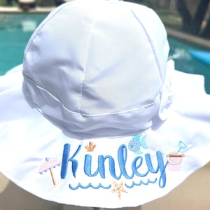 May include: White sun hat with a wide brim, embroidered with the name "Kinley" in blue script. The design includes beach-themed elements like an umbrella, seashell, starfish, and sand pail.