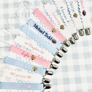 May include: A collection of personalized baby pacifier clips with embroidered names and designs. The clips are made of white satin ribbon with scalloped edges and feature a variety of designs, including a football, a dog, and a baby carriage. The clips are attached to a silver metal clip.