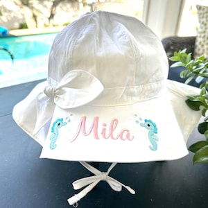 May include: White sun hat with a pink embroidered name "Mila" and two blue seahorse designs on the brim.