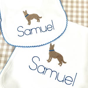 May include: Two white cotton burp cloths with blue trim. One burp cloth has a brown embroidered dog and the name "Samuel" embroidered in blue. The other burp cloth has the same design.
