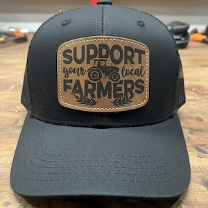 May include: Black trucker hat with a curved brim and mesh back. A brown leather patch on the front displays the text "SUPPORT your LOCAL FARMERS" with a tractor graphic. The hat is designed for outdoor wear and supports local agriculture.