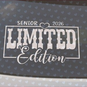 May include: White decal on a car window with the words "SENIOR 2026 LIMITED Edition." The design includes a heart symbol. The background is a dark gray with a pattern of small white hearts.
