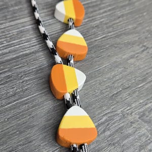 Könnte beinhalten: Eine Halskette mit vier candy-corn-förmigen Perlen, die auf einer schwarz-weiß gemusterten Schnur mit Hahnentrittmuster aufgereiht sind. Jede Perle ist mit horizontalen Streifen in Weiß, Gelb und Orange bemalt, die das Aussehen von Candy Corn nachahmen.