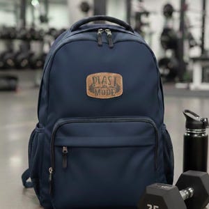 May include: Navy blue backpack with a front pocket and side mesh pocket. The backpack has a leather patch with the text "PLAST MODE". A black water bottle and a 25-pound dumbbell are next to the backpack.