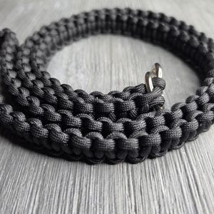 May include: A black paracord leash featuring a tightly woven, textured knot pattern throughout the length of the cord. The leash includes a polished silver-toned metal ring attachment at one end, providing a secure point for connection.