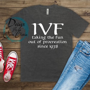 Puede incluir: Camiseta gris oscuro con cuello en V y texto blanco "IVF taking the fun out of procreation since 1978." La camiseta se combina con vaqueros azules y zapatillas rojas con cordones blancos.