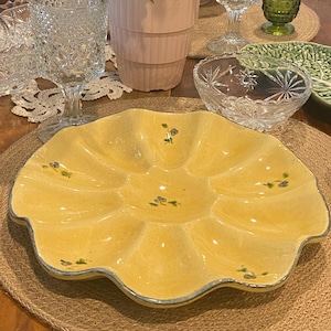 May include: A yellow ceramic serving dish with multiple compartments, adorned with small floral accents. The dish is surrounded by glassware, including clear goblets and a green tumbler, and a potted fern. The items are arranged on a woven placemat.