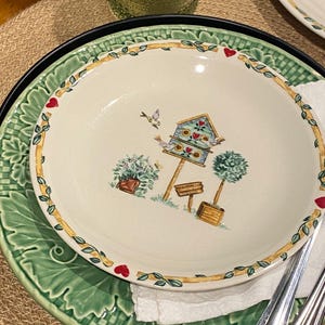 May include: A round ceramic plate with a cream-colored center featuring a colorful illustration of a birdhouse, two birds, a potted plant, and a topiary. The rim is decorated with a yellow border, green leaves, and small red heart accents.