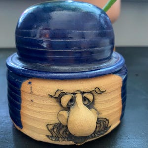 May include: A ceramic jar with a lid shaped like a dark blue bowler hat. The base is unglazed tan clay featuring a sculpted face with a prominent nose, etched eyes, and a mustache. The sides of the jar are finished in a glossy dark blue glaze.