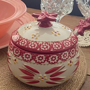 May include: A white ceramic sugar bowl with a red floral design and a matching lid. The lid is topped with a red flower-shaped handle. The bowl has a red and white patterned border and is decorated with red and yellow floral accents.