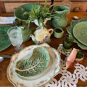 May include: A collection of green cabbage-themed ceramic dinnerware including plates, a pitcher, and a bowl, alongside a cottage-shaped teapot, a pink bird figurine, clear and green textured glassware, and a white lace doily on a wooden table.