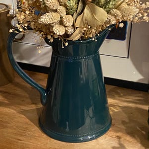 May include: A teal-colored ceramic pitcher with a handle, filled with dried flowers and foliage. The pitcher has a glossy finish and a decorative beaded rim. The arrangement includes beige, brown, and green elements, creating a rustic, natural aesthetic.