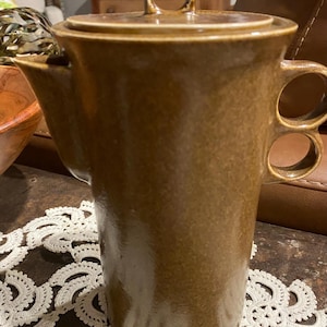 May include: A brown ceramic coffee pot with a lid and a handle. The pot has a spout and two finger holes on the handle. The pot is sitting on a decorative white lace doily.